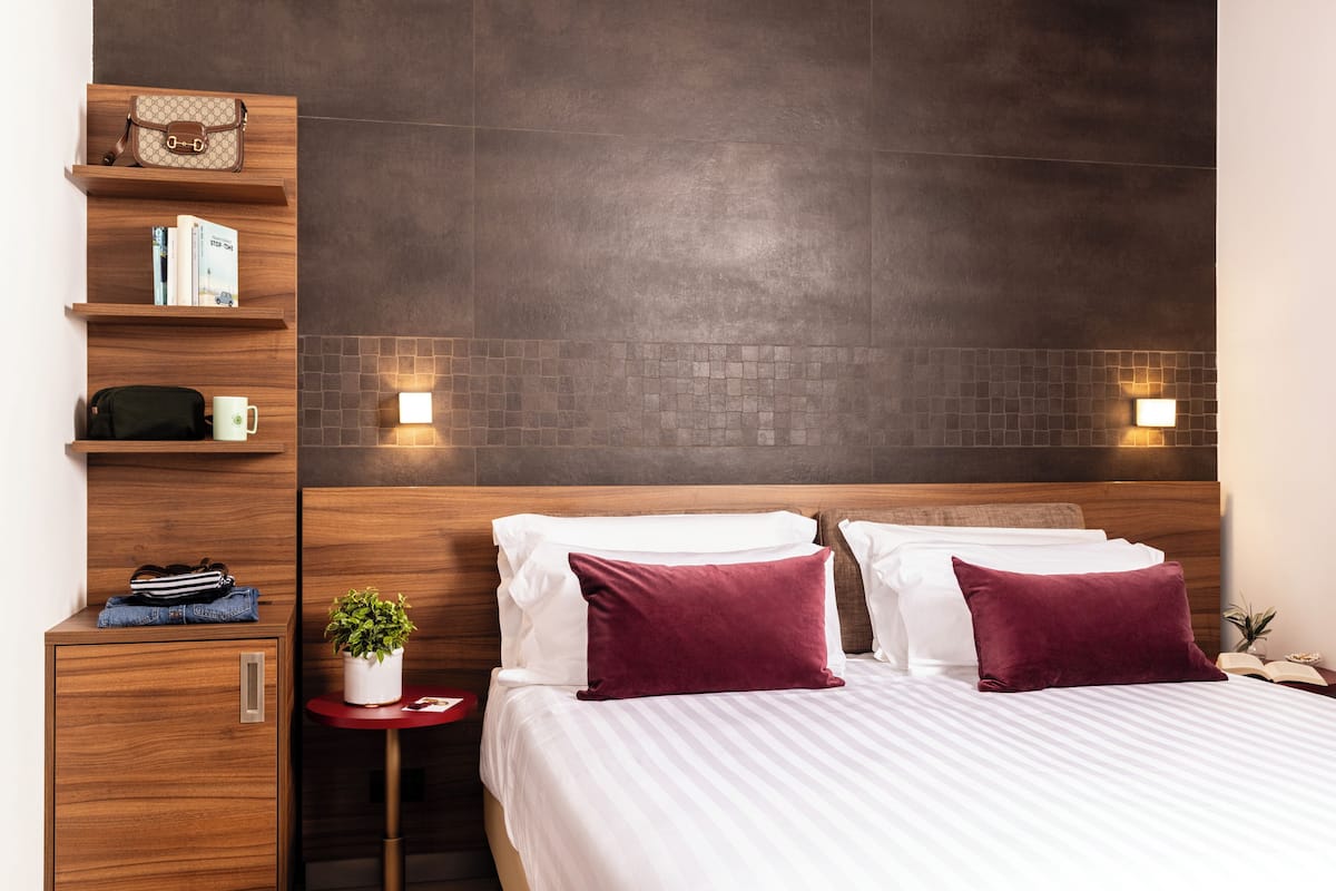 A hotel room with a wooden headboard, a bed with white and maroon pillows, a small side table with a plant, and a shelf with books and a handbag.