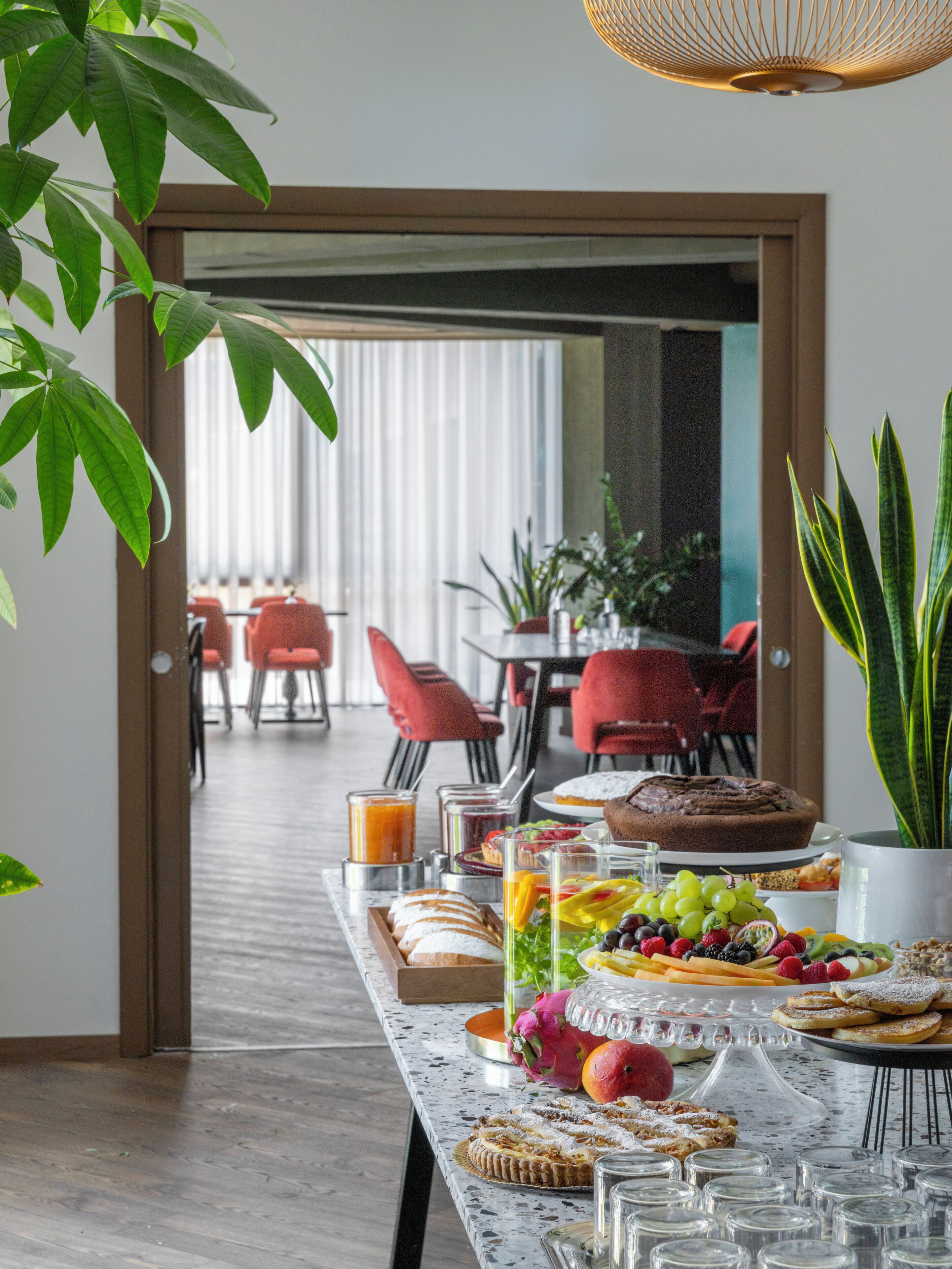 A breakfast buffet with various food items and a view of the dining area with red chairs and a table.