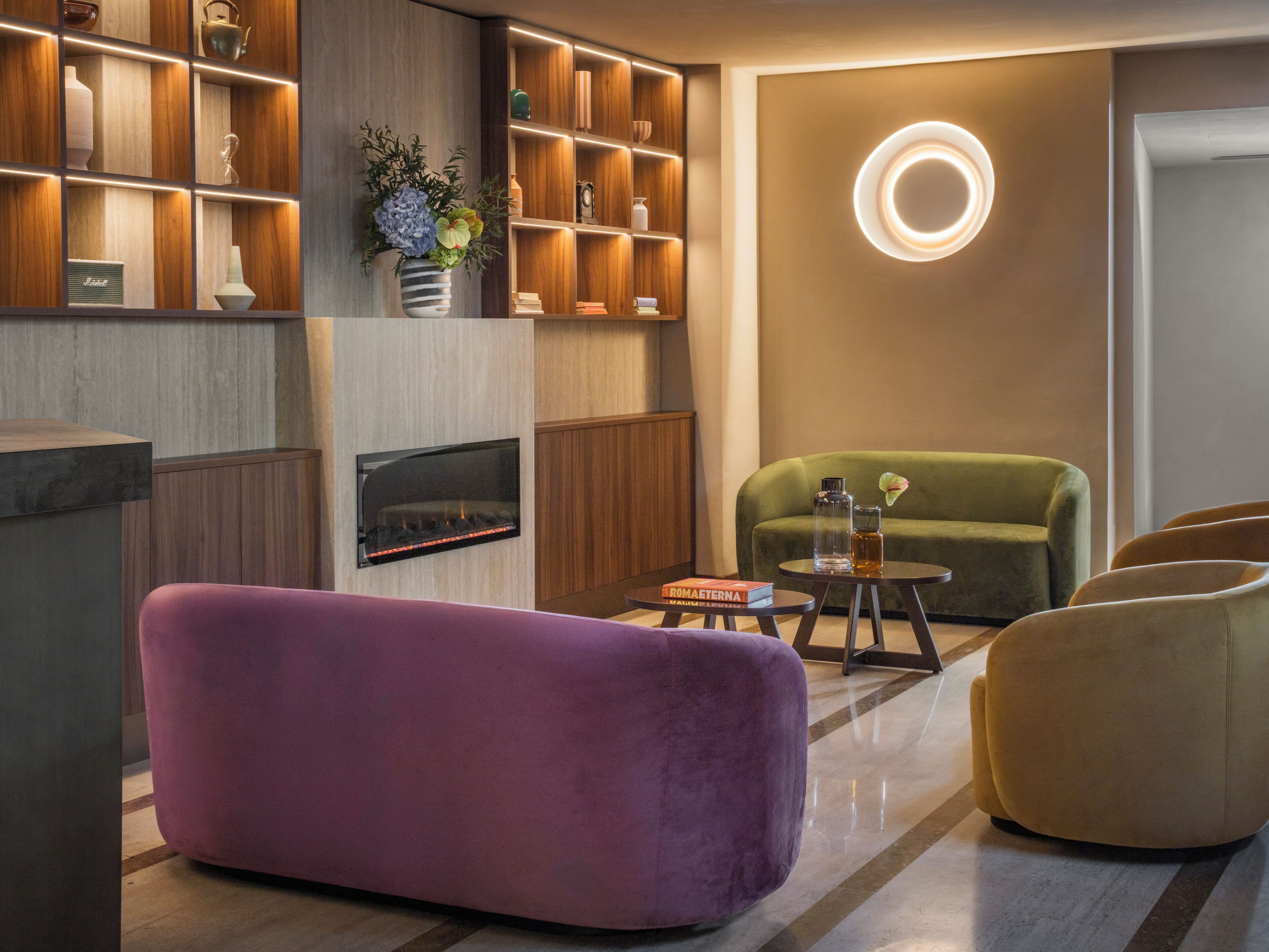 Een moderne woonkamer met een open haard, een groene bank, een paars voetenbankje en een houten boekenkast.
