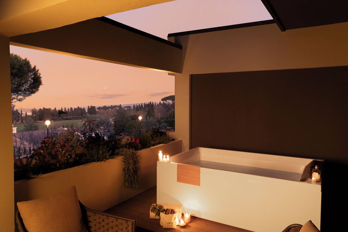 A modern bathroom with a bathtub, candles, and a view of a garden at dusk.