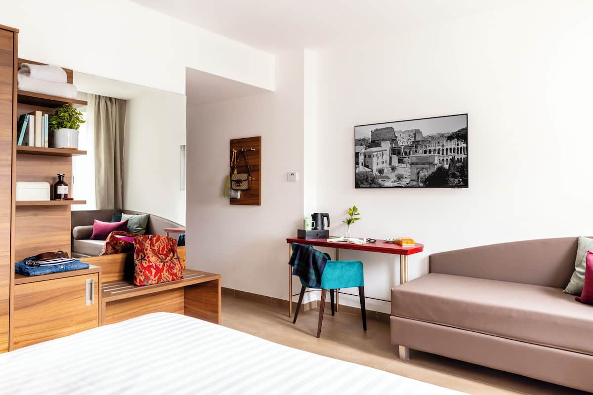A modern hotel room with a bed, a sofa, a desk with a chair, a bookshelf, and a framed picture on the wall.