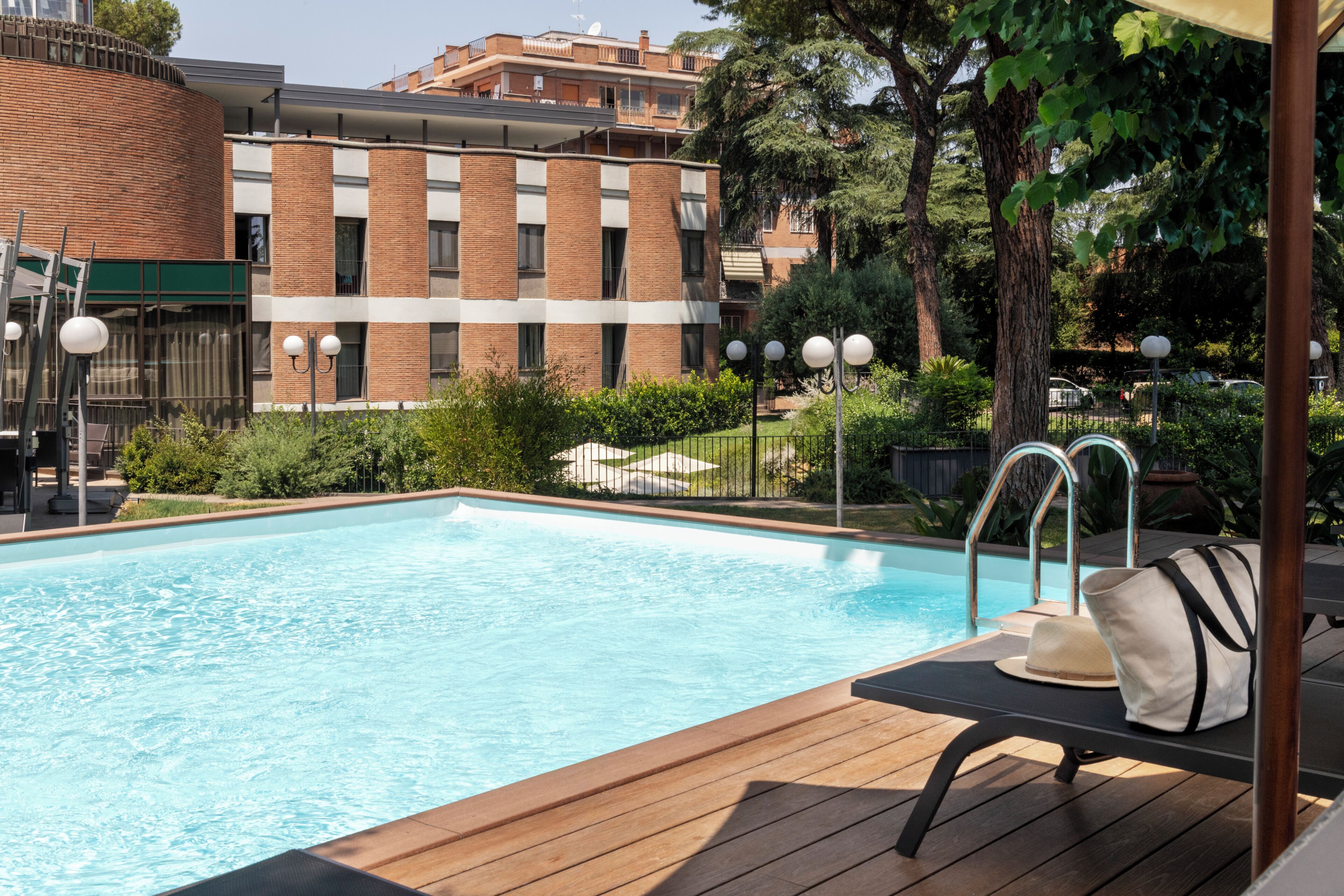 Una piscina con un solarium in legno, una sedia a sdraio e vista su un edificio in mattoni con balconi.