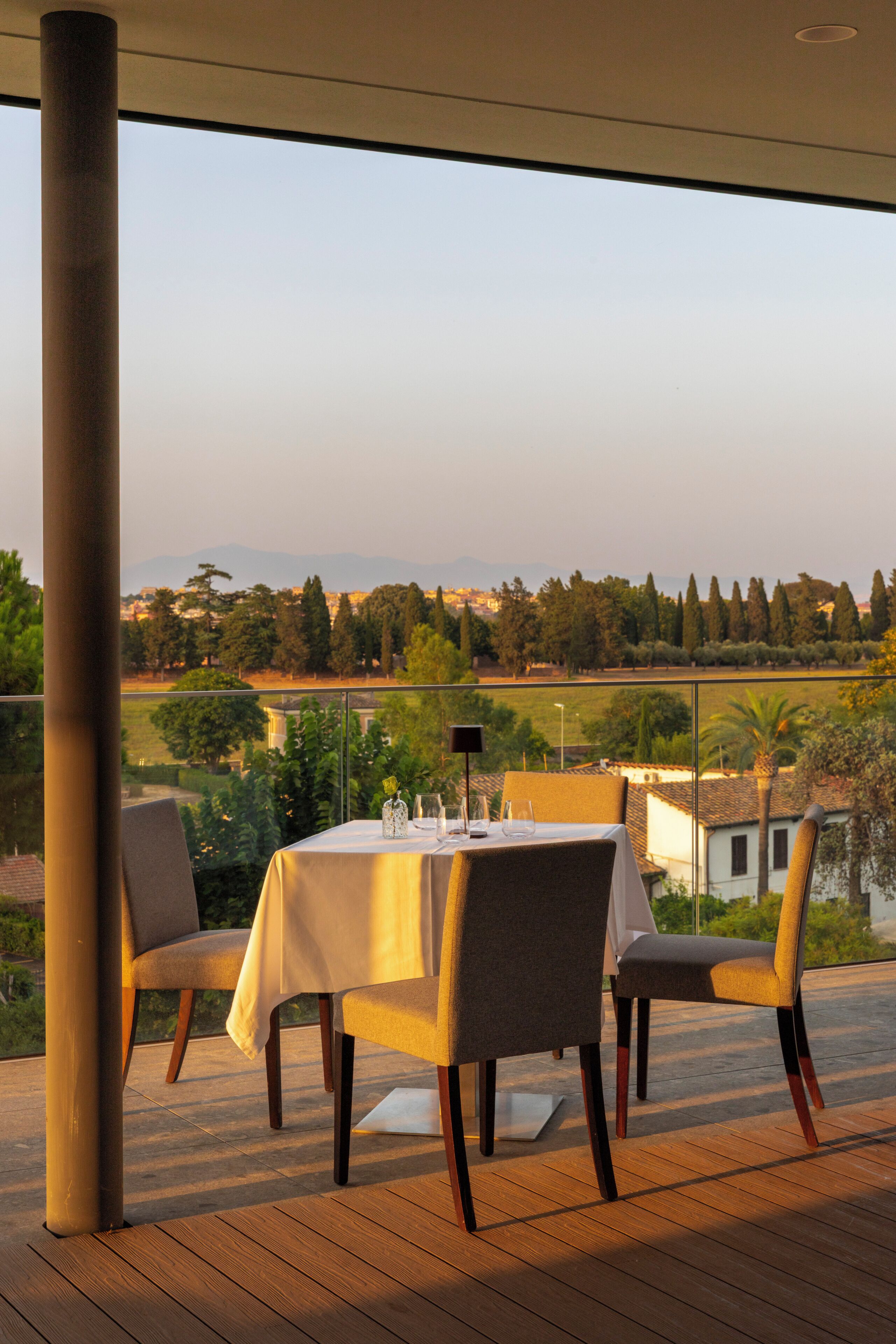 A dining table with chairs set for two, overlooking a scenic landscape with trees and buildings.