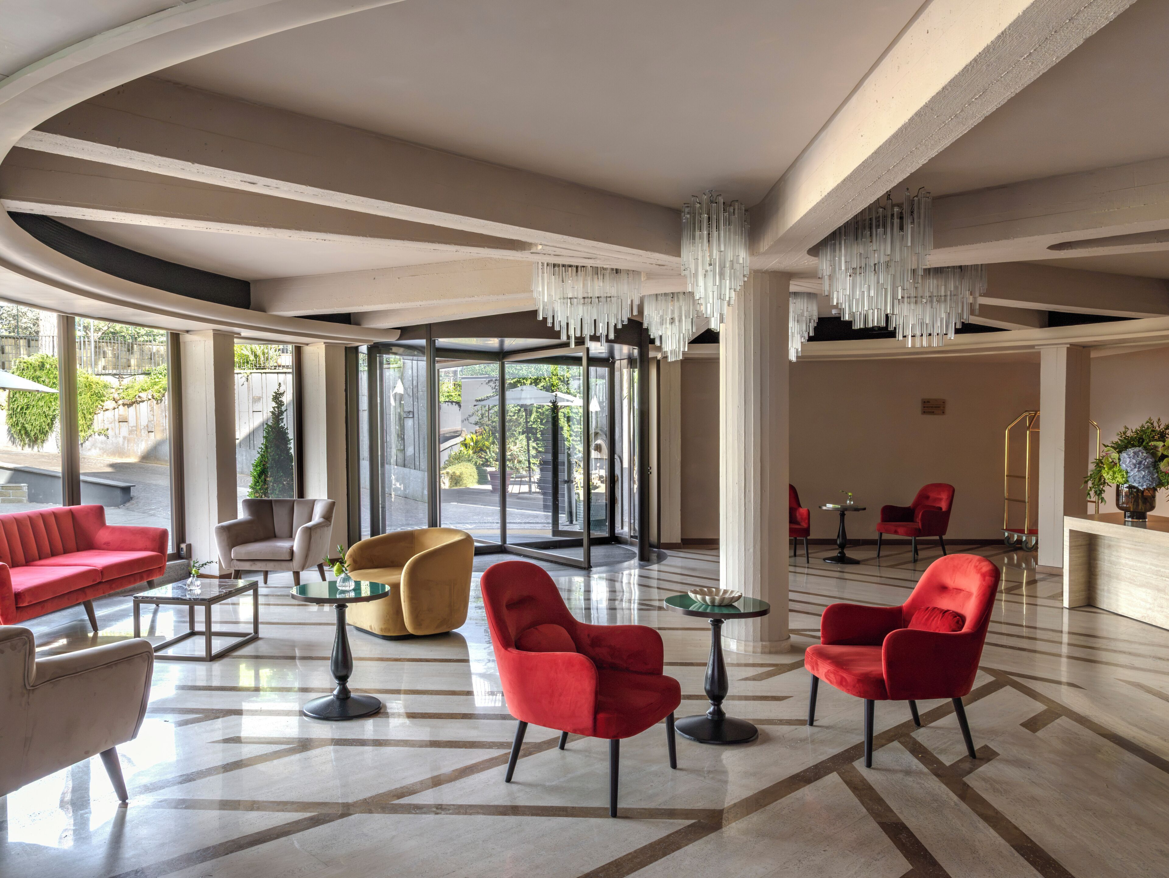 Un vestíbulo amplio con diseño moderno, que cuenta con un sofá rojo, un sillón amarillo y sillas rojas, con un gran ventanal que da al exterior.