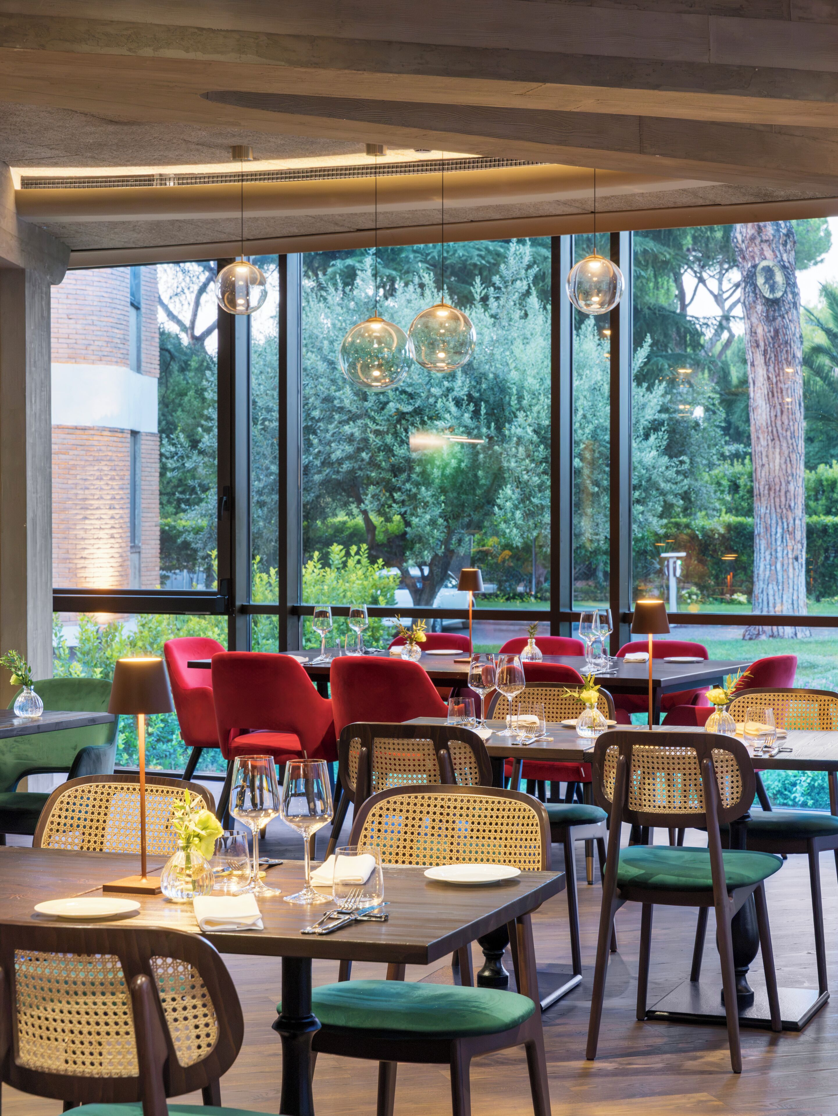 A restaurant with tables set for dining, red chairs, and a view of greenery outside.