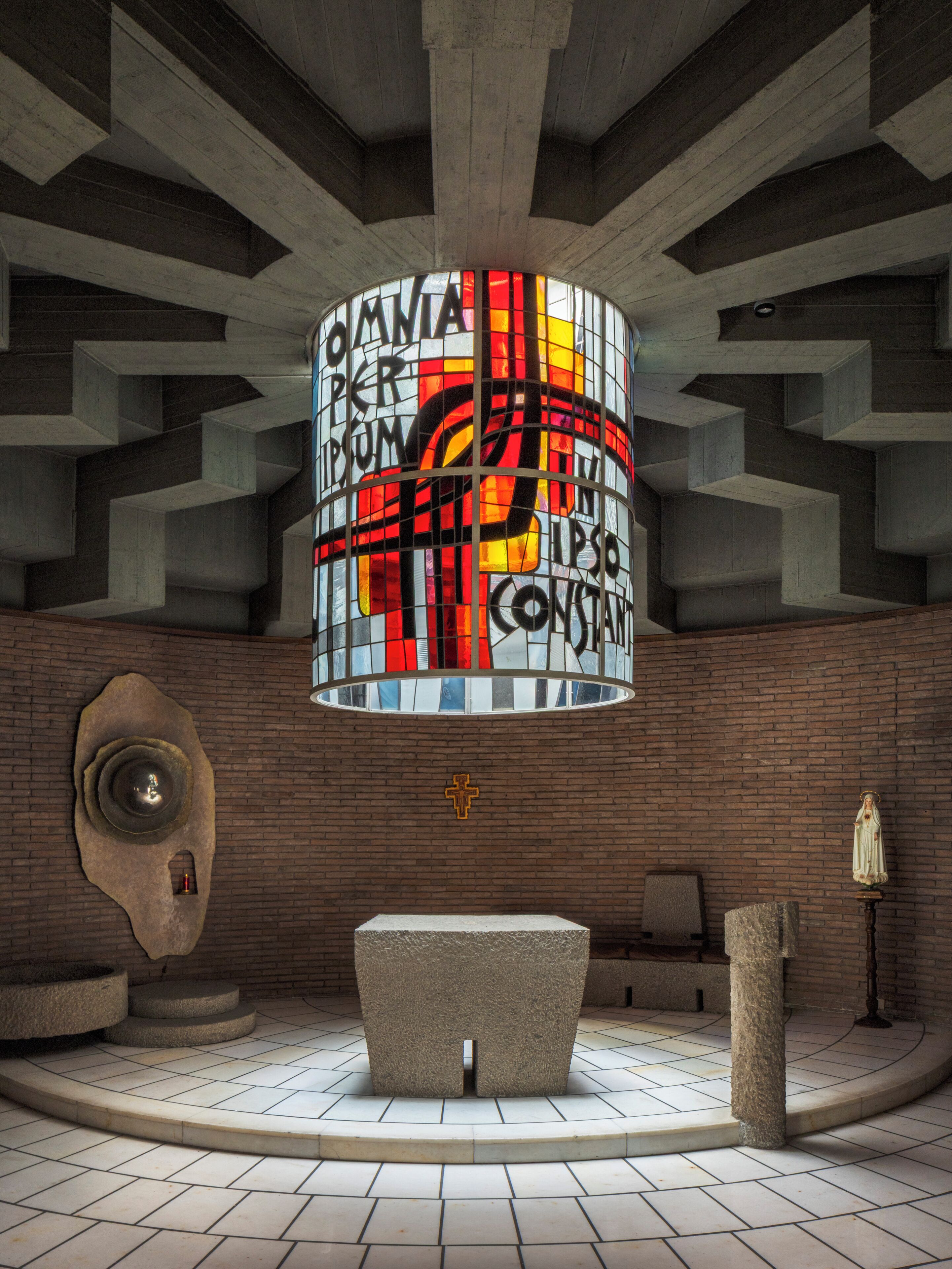 A church interior with a central stained glass window, a brick wall, a stone altar, and a statue on a pedestal.