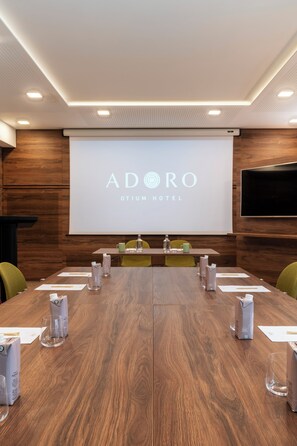 A conference room with a long table, chairs, a projector screen displaying 'ADORO OTIUM HOTEL', and a wooden wall.