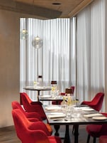 Zona pranzo con tavolo imbandito per quattro persone, sedie rosse rivestite in tessuto e un'ampia finestra con persiane verticali.