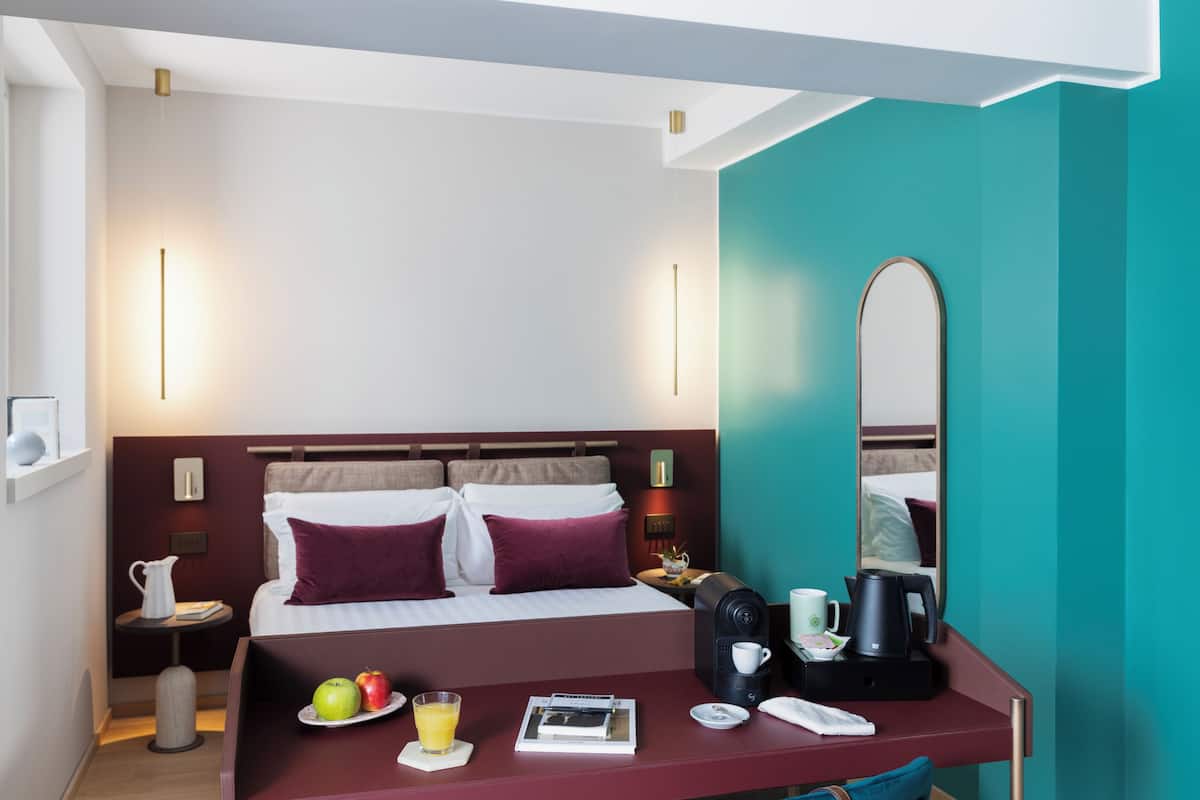 A hotel room with a bed, bedside table, and a desk with a chair. There is a mirror on the wall, a coffee maker, and a fruit plate with apples and a glass of orange juice.