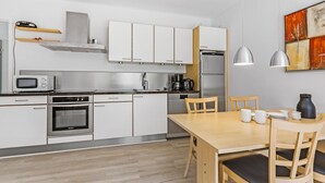Een moderne keuken met witte kasten, roestvrijstalen apparatuur, een houten eettafel met stoelen, en een schilderij aan de muur.