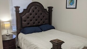 Une chambre avec un grand lit, une table de chevet, une lampe, un ventilateur de plafond, une fenêtre avec des stores et un tableau encadré au mur.