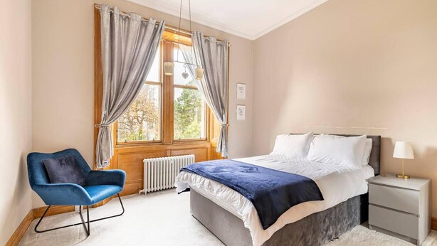A bedroom with a bed, a chair, a nightstand, and a window with curtains. - Stunning 5 Bedroom Large Apartment With Parking (Edinburgh)