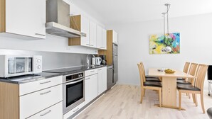 Eine moderne Küche mit weißen Schränken, einer Mikrowelle, einem Backofen und einer Kaffeemaschine. Es gibt einen Essbereich mit einem Holztisch und Stühlen.