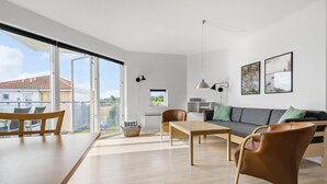 Een moderne woonkamer met een houten eettafel, een bank en een salontafel. Er zijn twee stoelen, een hanglamp en een raam met uitzicht op het omliggende landschap.