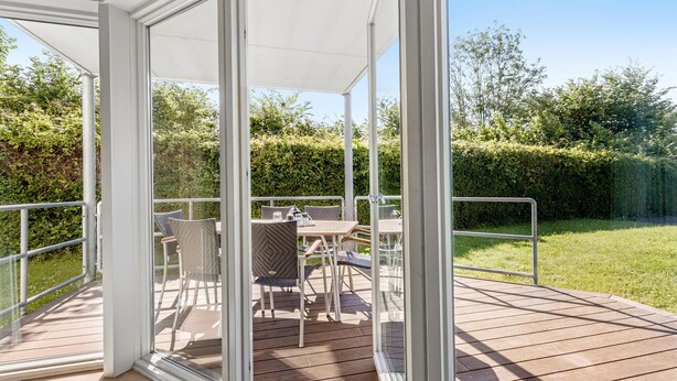 Eine überdachte Terrasse mit Tisch und Stühlen, umgeben von einem Metallgeländer und Blick auf einen grünen Garten.