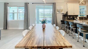 A spacious dining area with a wooden table, white chairs, and a view of a body of water through large windows. - Storey Lake Amazing Water view, Game Room, Movie Room and Theme Rooms (Kissimmee)