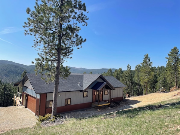 A modern cabin with a sloped roof, large windows, and a covered porch. It is surrounded by a forested landscape with mountains in the background. - Spacious, 3 bedroom private cabin on 6 acres. Near Mickelson trail & Deadwood (Lead)