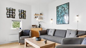 Ein modernes Wohnzimmer mit einem grauen Sofa, einem Holztisch und einem grauen Sessel. Ein Radio steht auf einer Holzkommode, eine Lampe ist vorhanden, und eine Wandablage zeigt dekorative Gegenstände.