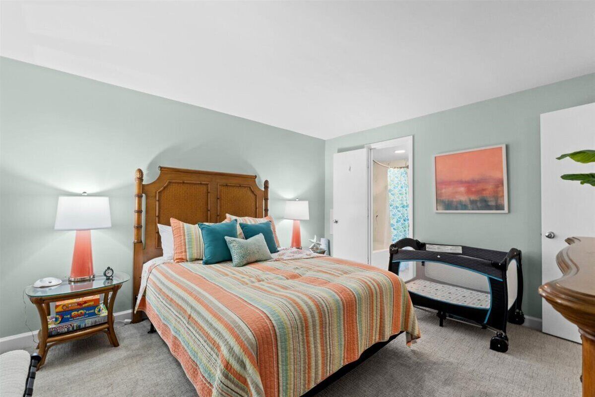 A bedroom with a wooden bed, striped bedding, a bedside table with books, a nightstand, a crib, and a door leading to another room.