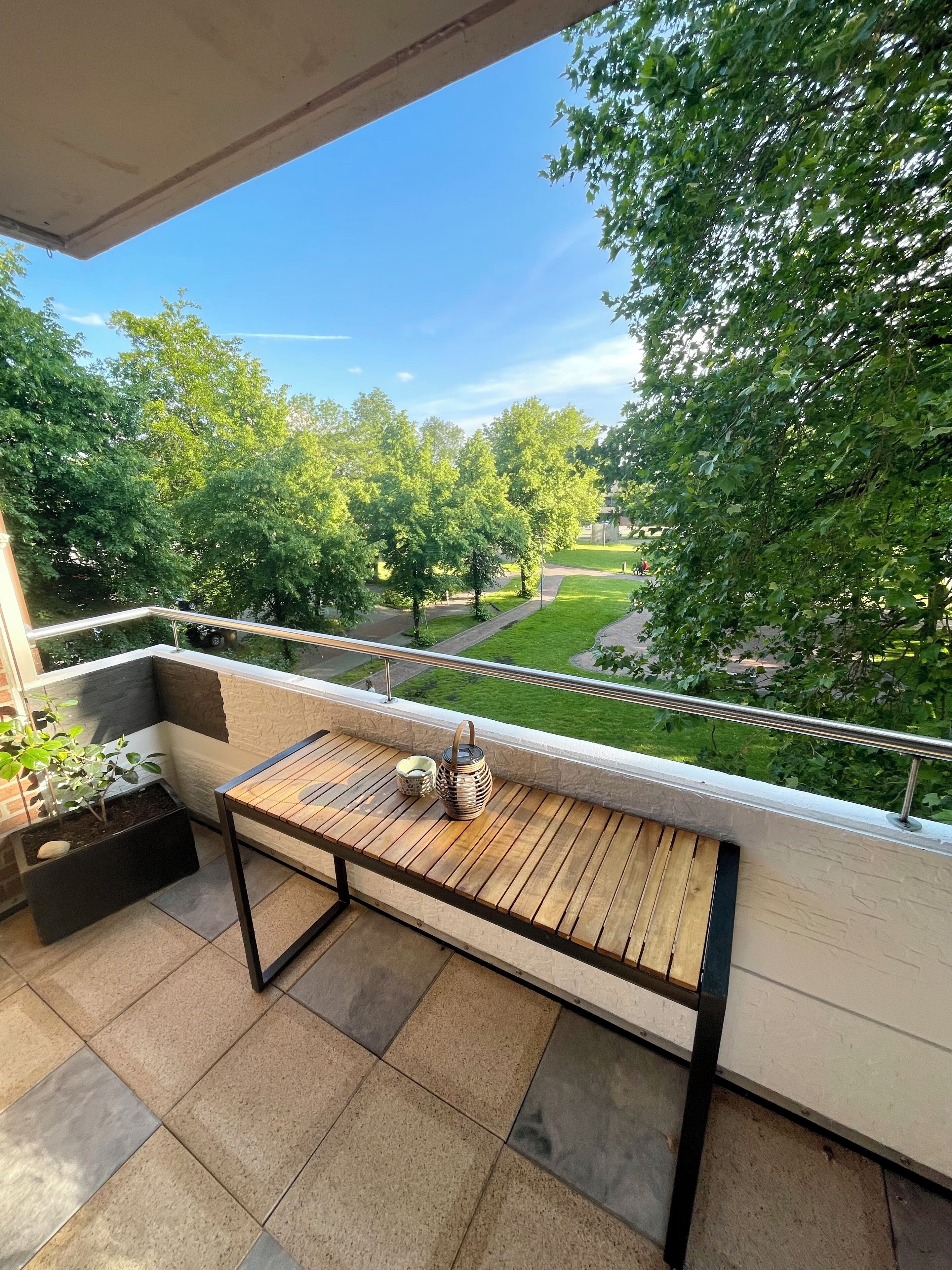 A balcony with a wooden table, a metal railing, and a view of a green park with trees and a lawn.