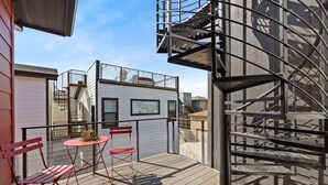 Un pontile in legno con sedie rosse e un tavolino, una scala a chiocciola in metallo e un edificio bianco con una finestra.