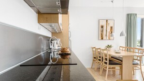 A modern kitchen with a black countertop, stainless steel appliances, and a wooden dining area with a table and chairs. - Gudhjem Søpark Lejl. 38 (Gudhjem)