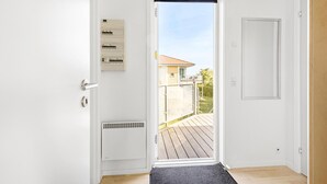 A white door with a black doormat, a wall-mounted electrical panel, and a view of a balcony with a wooden deck and a building in the background. - Gudhjem Søpark Lejl. 38 (Gudhjem)