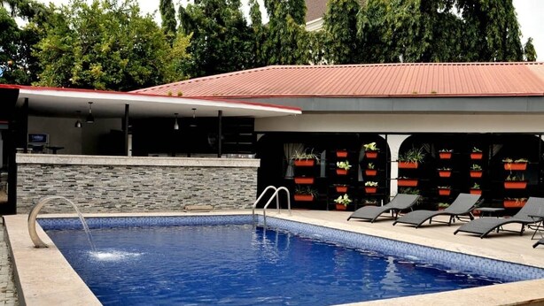 A rectangular swimming pool with a stone wall and a red roofed structure with outdoor seating and planters. - THE ADDRESS HOTEL AND TOWER (Abuja)