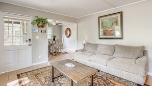 Un soggiorno con un divano color beige, un tavolino in legno, un tappeto decorato, una porta con finestra, una ventola a soffitto e un quadro incorniciato appeso al muro.