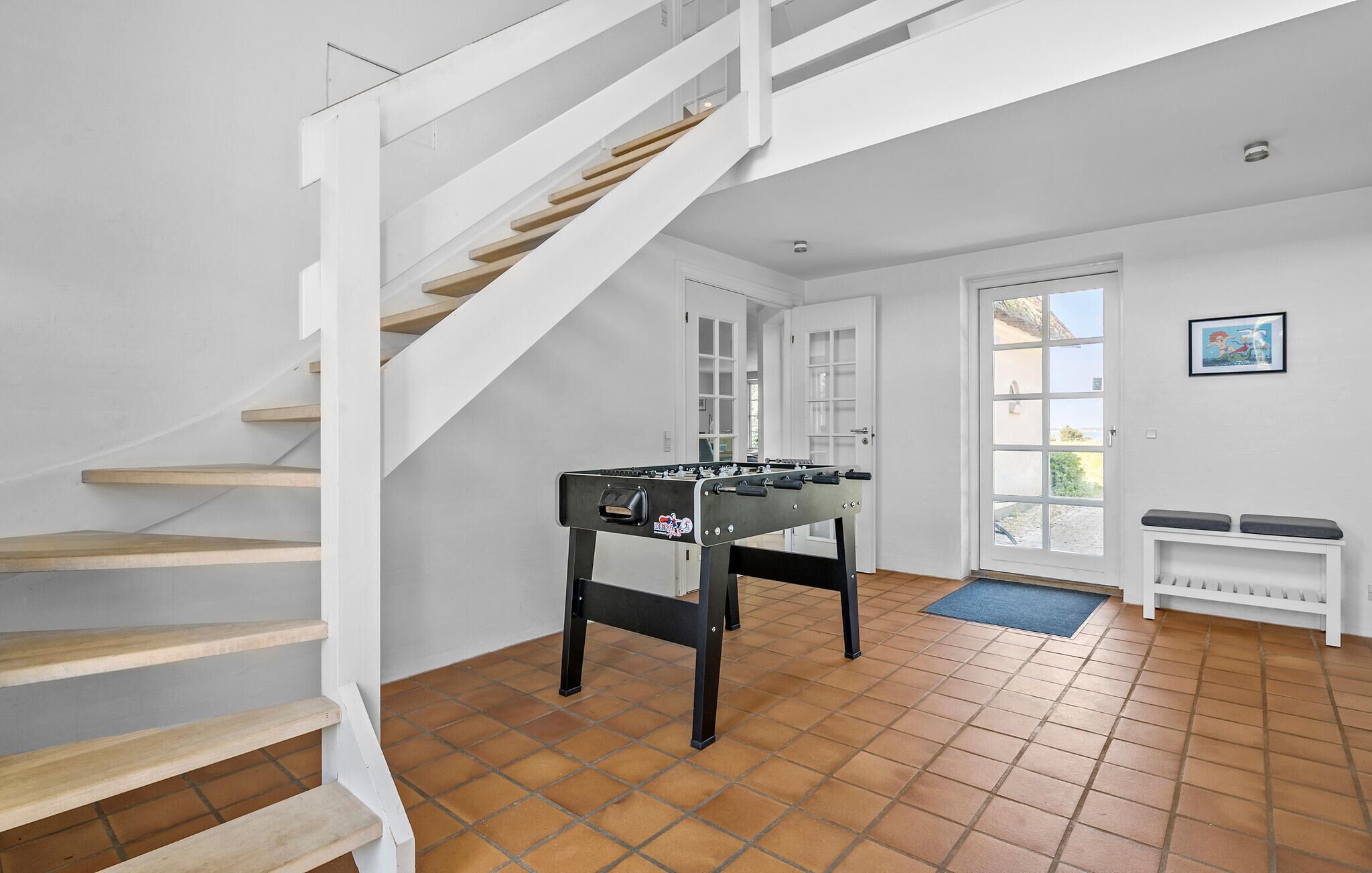 A spacious room with a wooden staircase, a foosball table, and a door leading outside.