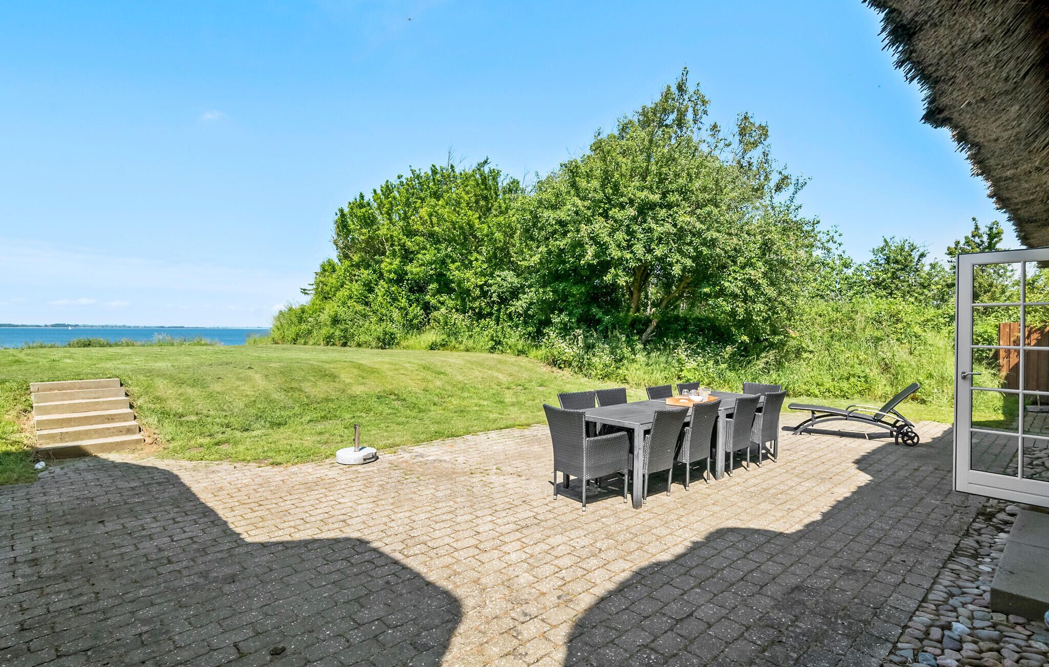 Een terras buiten met een tafel en stoelen, een constructie met rieten dak en uitzicht op zee en groen.