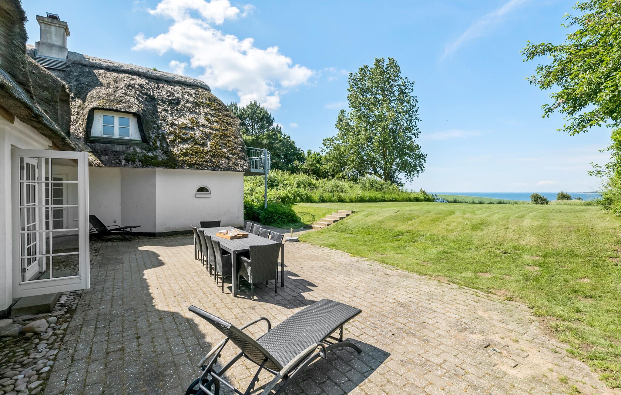 A thatched-roof house with a patio area featuring a table and chairs, a lounge chair, and a view of the sea.