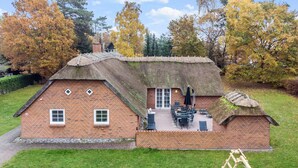 En stråtækt hytte med murstensfacade, en terrasse med udendørs møbler, og en stige i forgrunden.