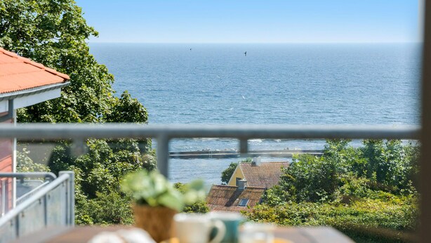 A view of the sea from a window overlooking a balcony with a plant, a table with a bowl, and a building with a red roof. - Gudhjem Søpark Lejl. 30 (Gudhjem)