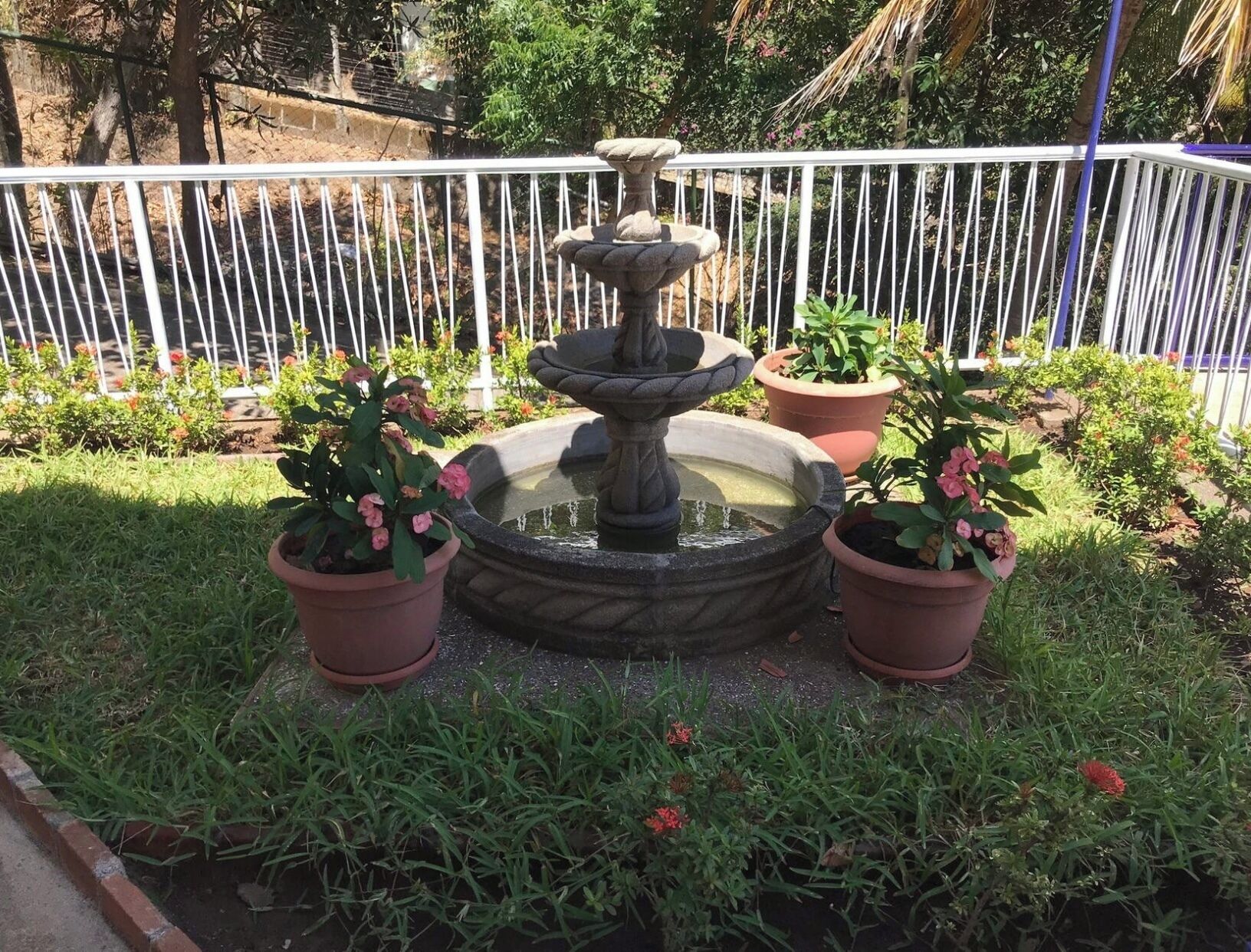 A multi-tiered stone fountain surrounded by potted plants and a white metal fence.