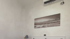 Un dormitorio con cama, almohadas, mesita de noche con lámpara, aire acondicionado de pared y un cuadro con una escena de playa.