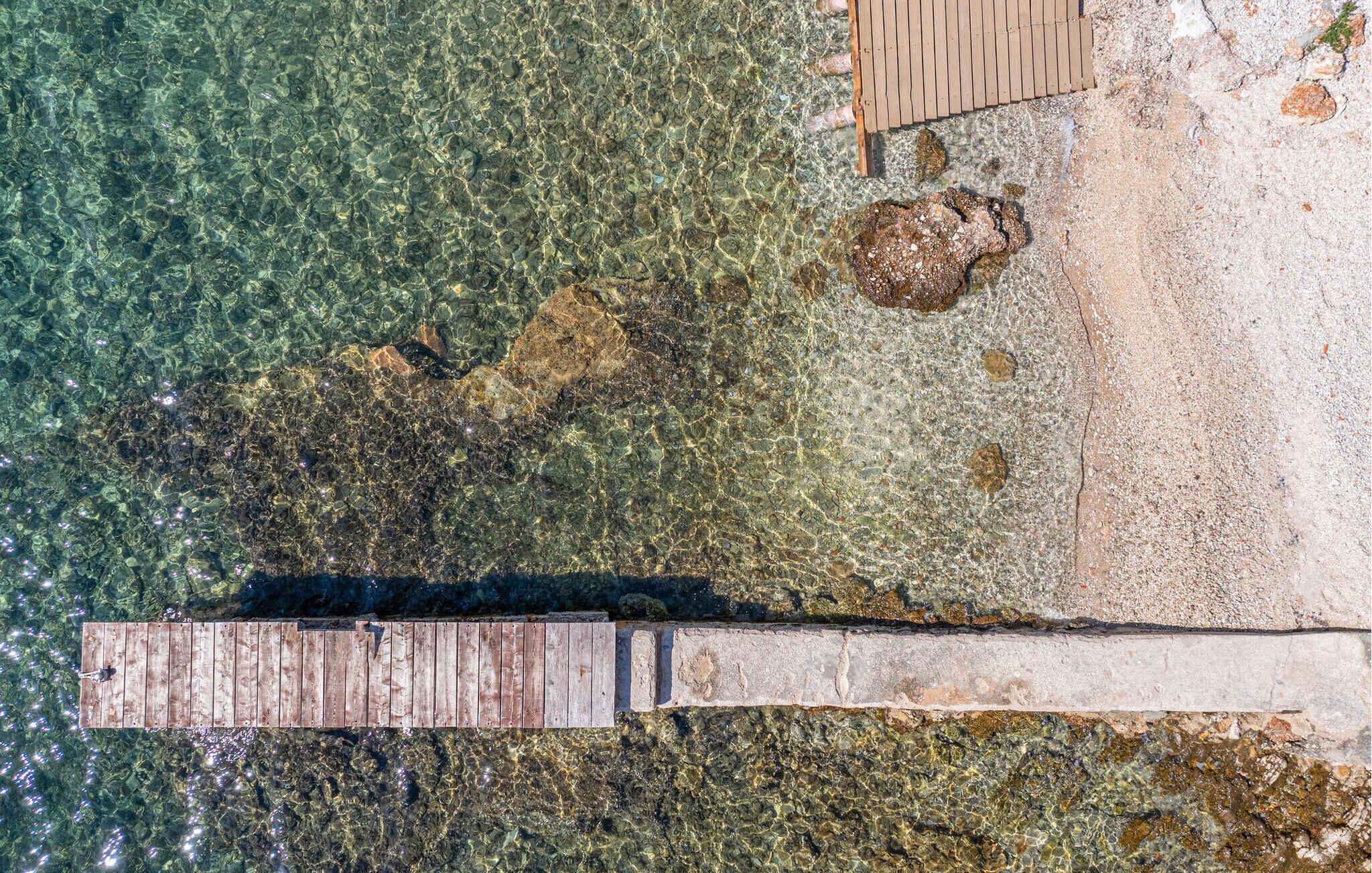 A wooden dock extending into clear shallow water, leading to a sandy beach with visible rocks and pebbles.