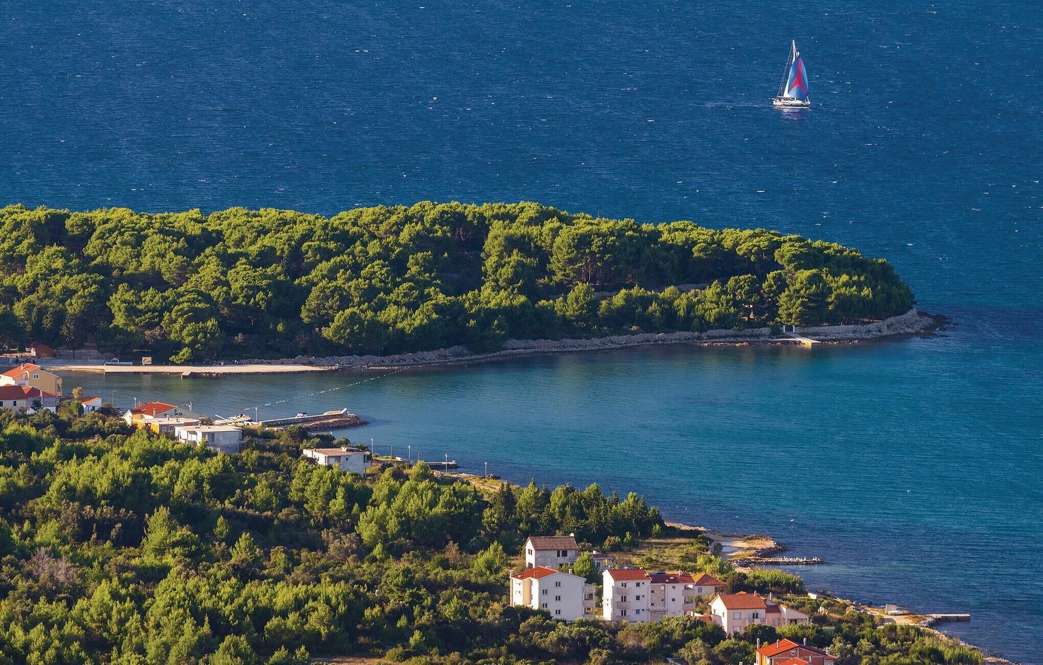 Una zona costiera con una piccola isola ricoperta da alberi verdi fitti, una barca a vela nell'acqua e un gruppo di edifici con tetti rossi vicino alla riva.