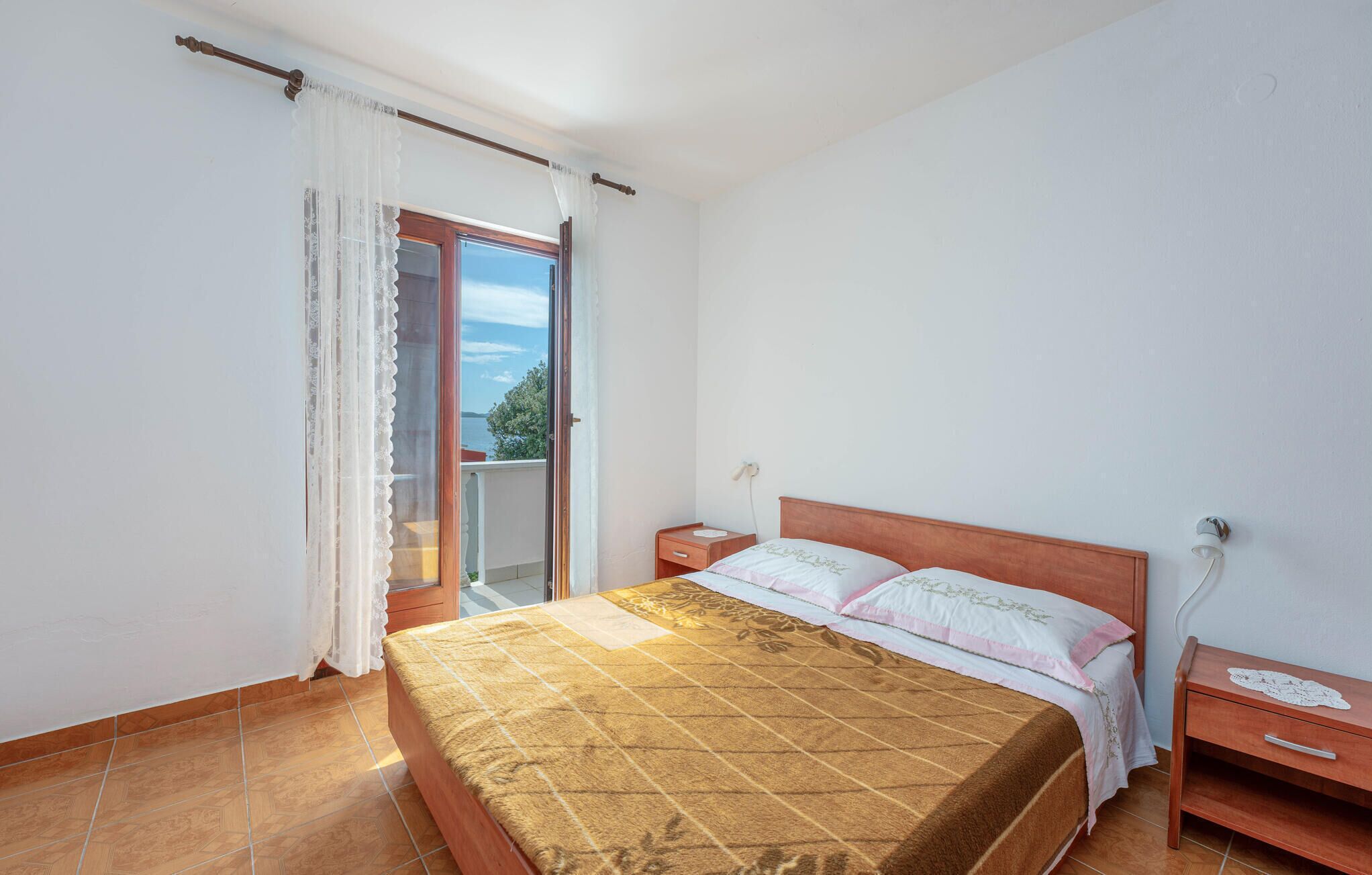Un dormitorio con cama, mesita de noche, cómoda de madera, ventana con cortinas y vistas al exterior.