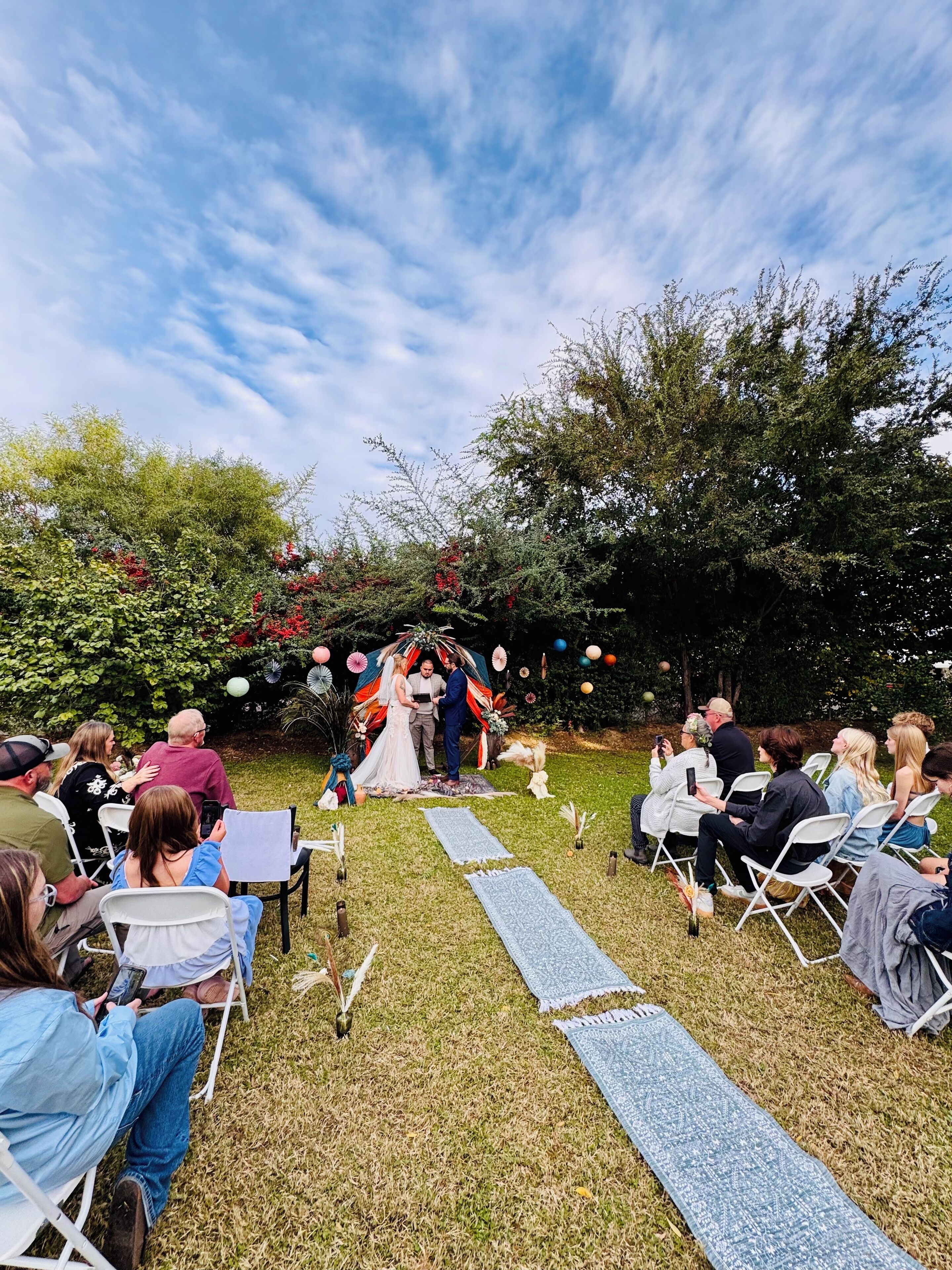 Una ceremonia de boda al aire libre con invitados sentados en sillas de jardín, un arco decorado y cielo despejado.