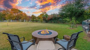 A backyard with a circular fire pit table surrounded by chairs, a green lawn, and trees with autumn foliage.