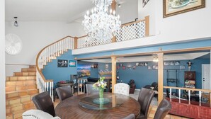 A spacious living area with a round wooden dining table surrounded by chairs, a chandelier, and a staircase leading to an upper level.