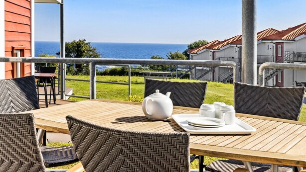 A wooden table with wicker chairs, a white teapot, and tea cups on a terrace overlooking the sea.