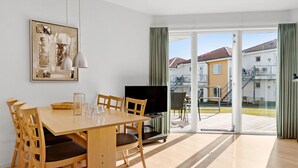 A modern living room with a wooden dining table, chairs, a flat-screen TV, and a large window with curtains.