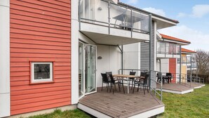 Una casa moderna de dos pisos con balcón, una terraza de madera y un área de comedor con mobiliario exterior.