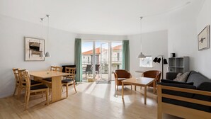 Una sala de estar moderna y bien iluminada con una mesa de comedor de madera, sillas, un sofá y un balcón con vistas a edificios.