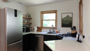 Una cocina moderna con un refrigerador, lavavajillas y estufa de acero inoxidable. Se aprecia una ventana con vista a zonas verdes, y un estante de madera con vajilla.
