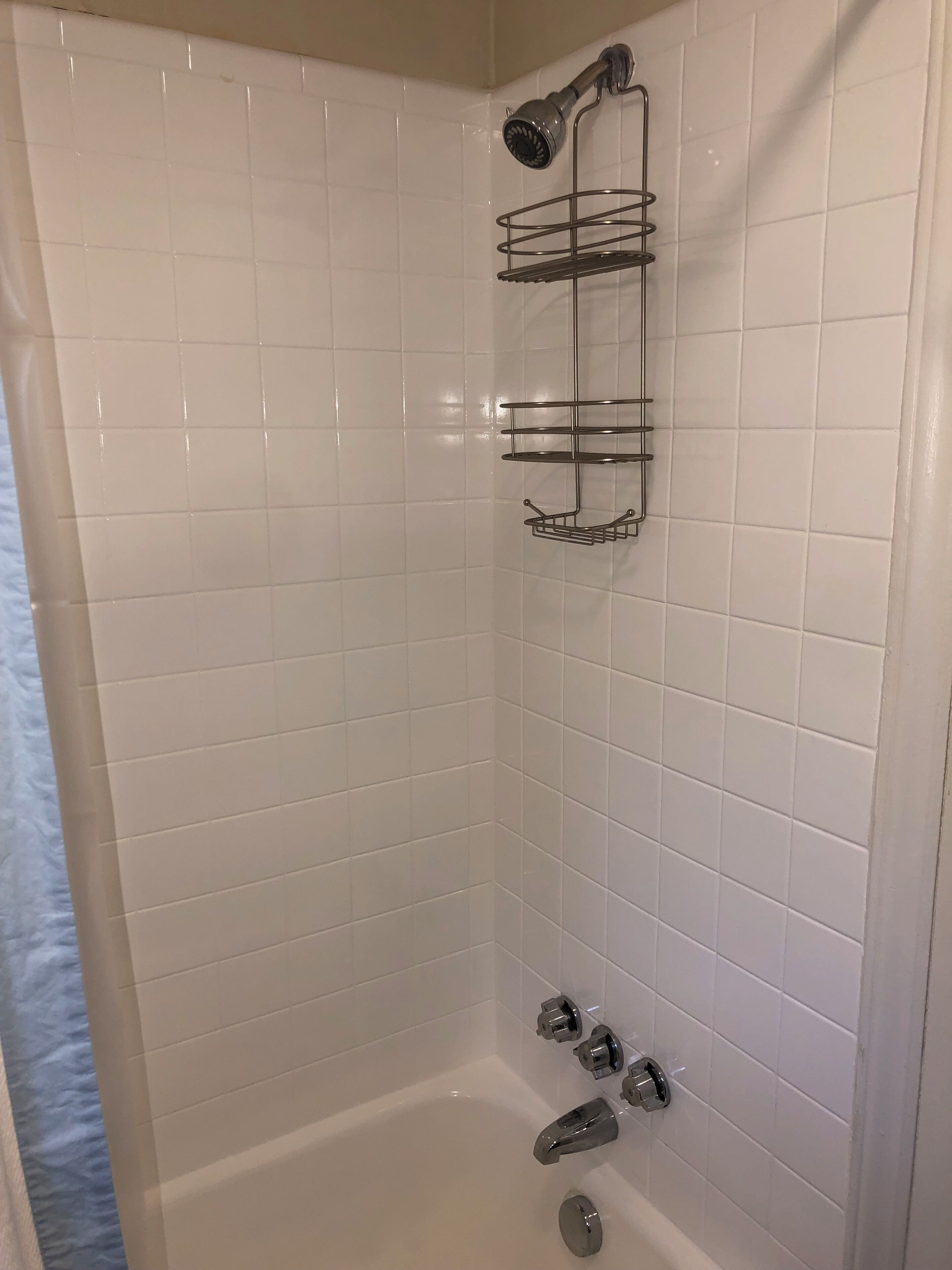 A shower area with white tiled walls, a showerhead, a metal shelf, and a bathtub with faucets.
