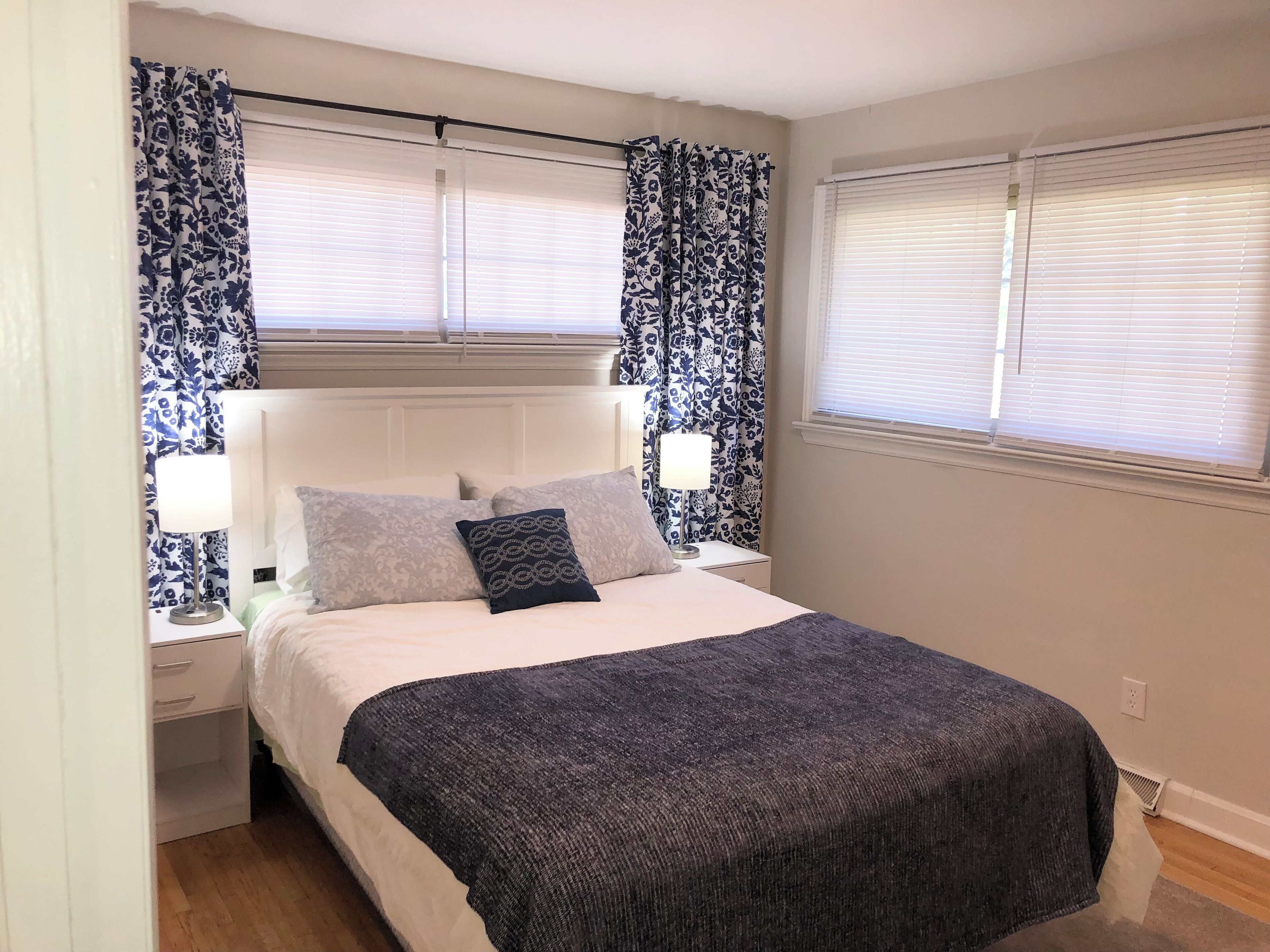 Een slaapkamer met een bed, twee nachtkastjes, een venster met zonwering en behangde gordijnen.