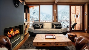 A cozy living room with a fireplace, a sofa with decorative pillows, a coffee table, and a view of snowy mountains through large windows. - Le Grand Capricia - mountain stay with sauna - OVO Network (La Clusaz)